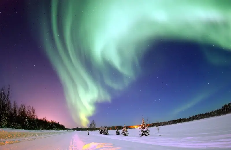 A huge green arc of northern lights over a snowy road and a lake edged with trees