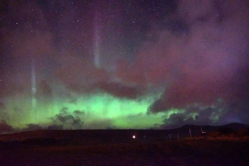 Green and pink northern lights low over a dark horizon, partly behind clouds