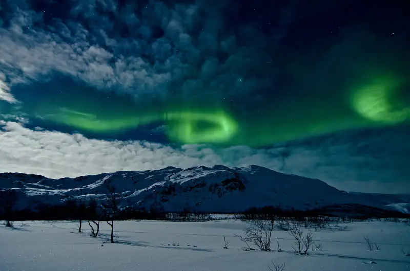 A green ring of northern lights over a snowy mountain under a cloudy, moonlit sky