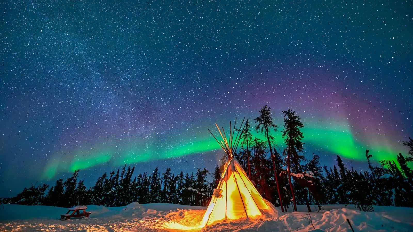 A tent glowing orange under green northern lights and the Milky Way, in a snowy forest