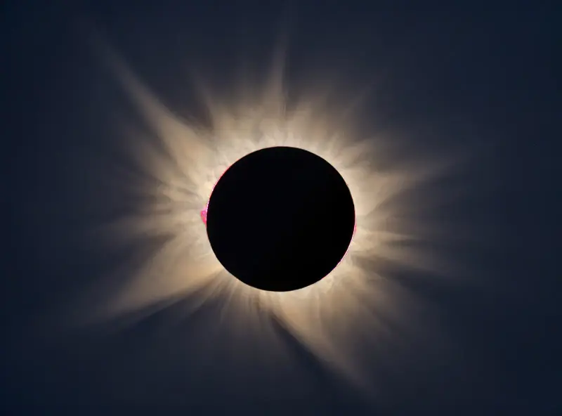 The white corona of the Sun around the black disc of the Moon during totality, against a deep blue sky