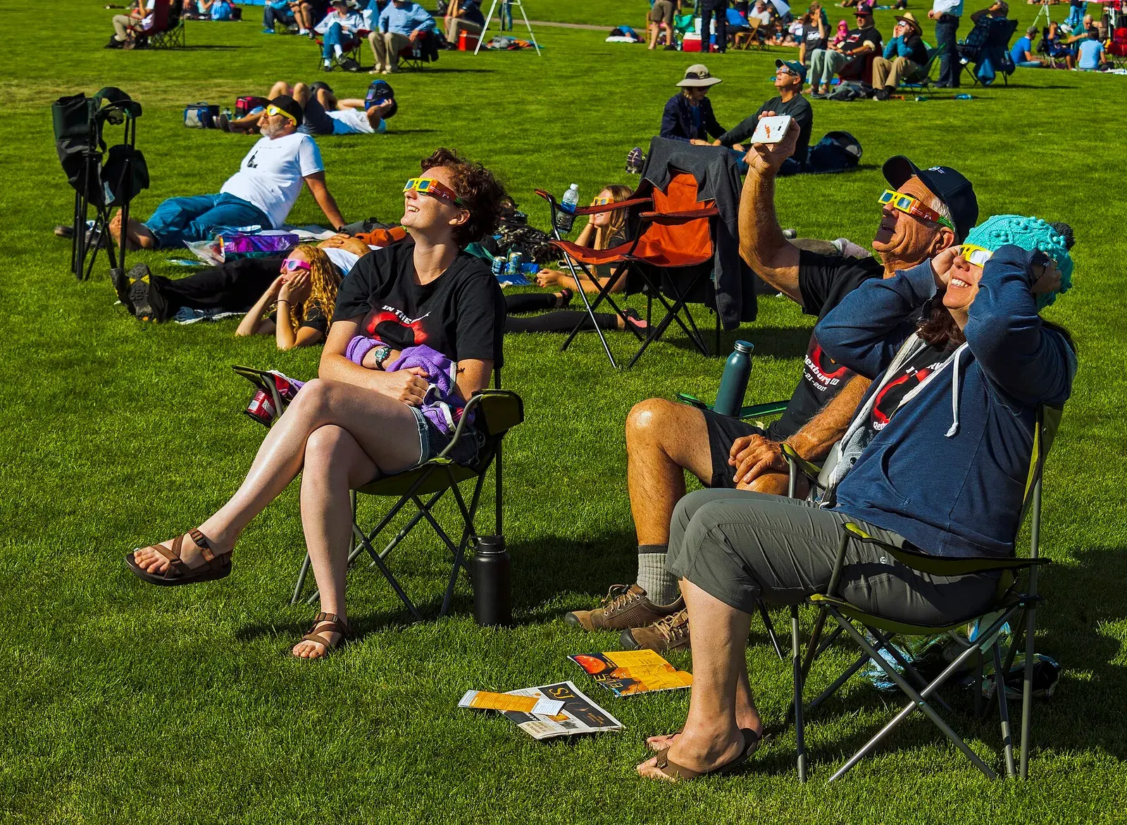 People sitting on folding chairs in a sunny park, looking up at the Sun through eclipse glasses
