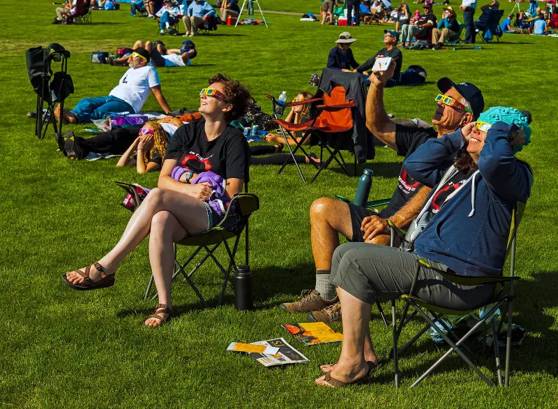 People sitting on folding chairs in a sunny park, looking up at the Sun through eclipse glasses