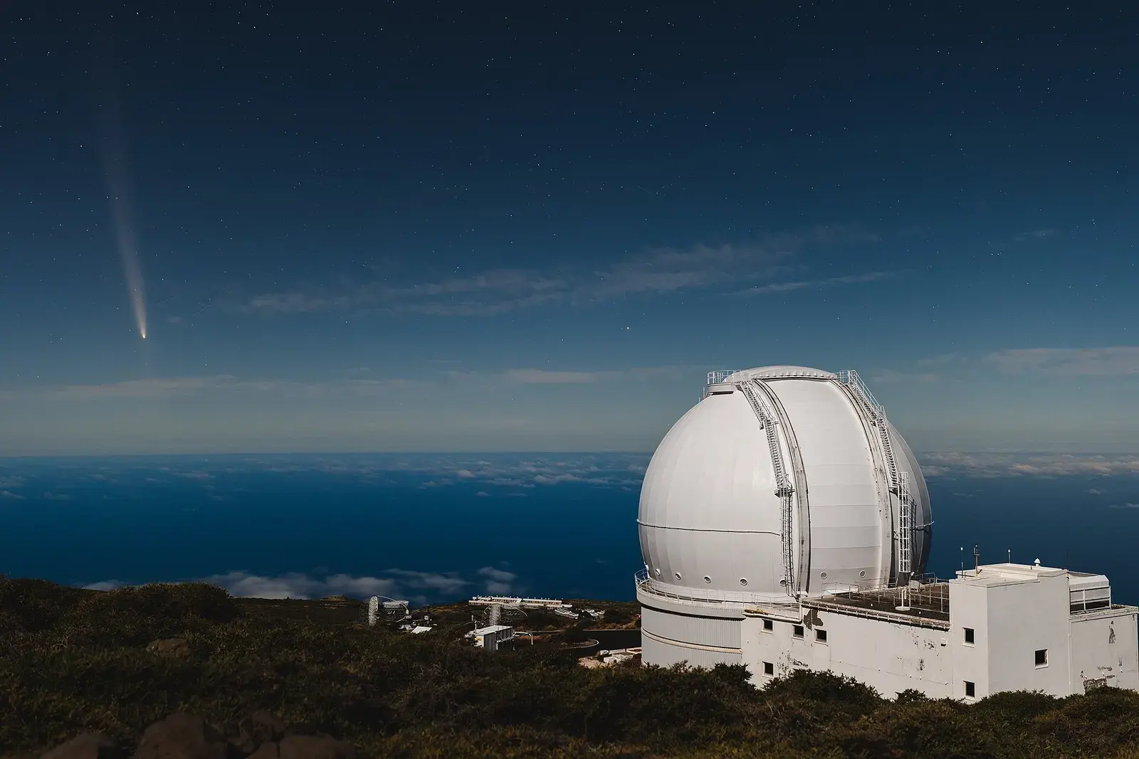The white dome of a large telescope on a mountain ridge above the sea, with a comet’s tail in the twilight sky