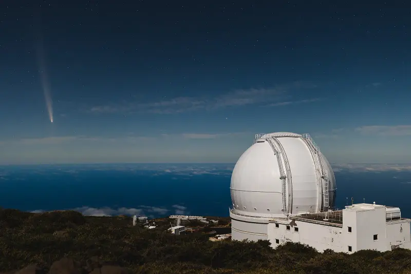 The white dome of a large telescope on a mountain ridge above the sea, with a comet’s tail in the twilight sky