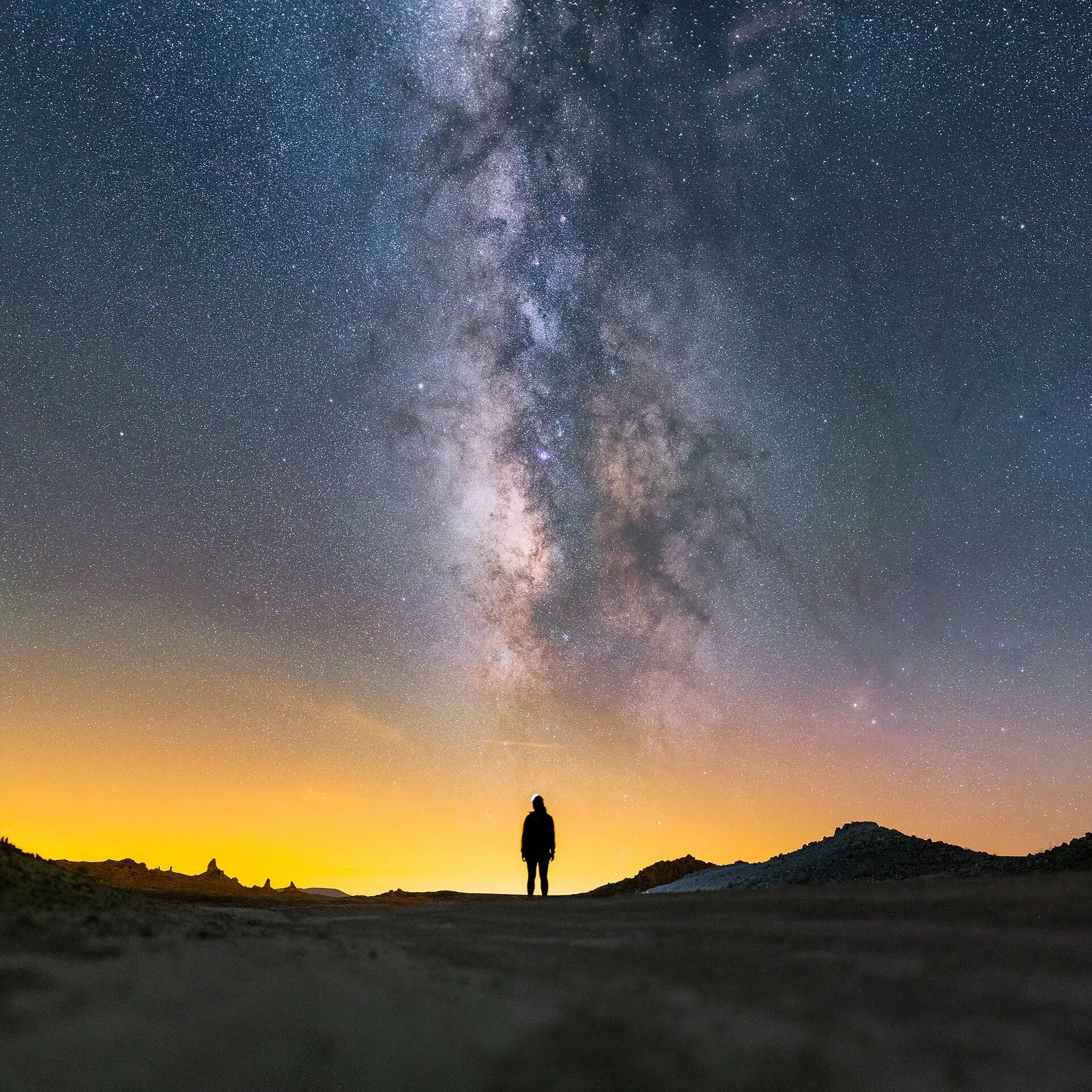 A person standing in silhouette under the Milky Way, with a glow of town lights on the horizon