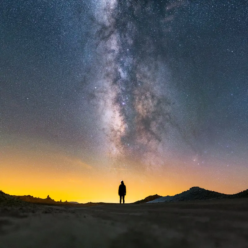 A person standing in silhouette under the Milky Way, with a glow of town lights on the horizon