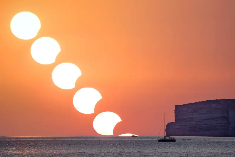 A sequence of the setting Sun with a growing bite taken out of it, over the sea next to a cliff