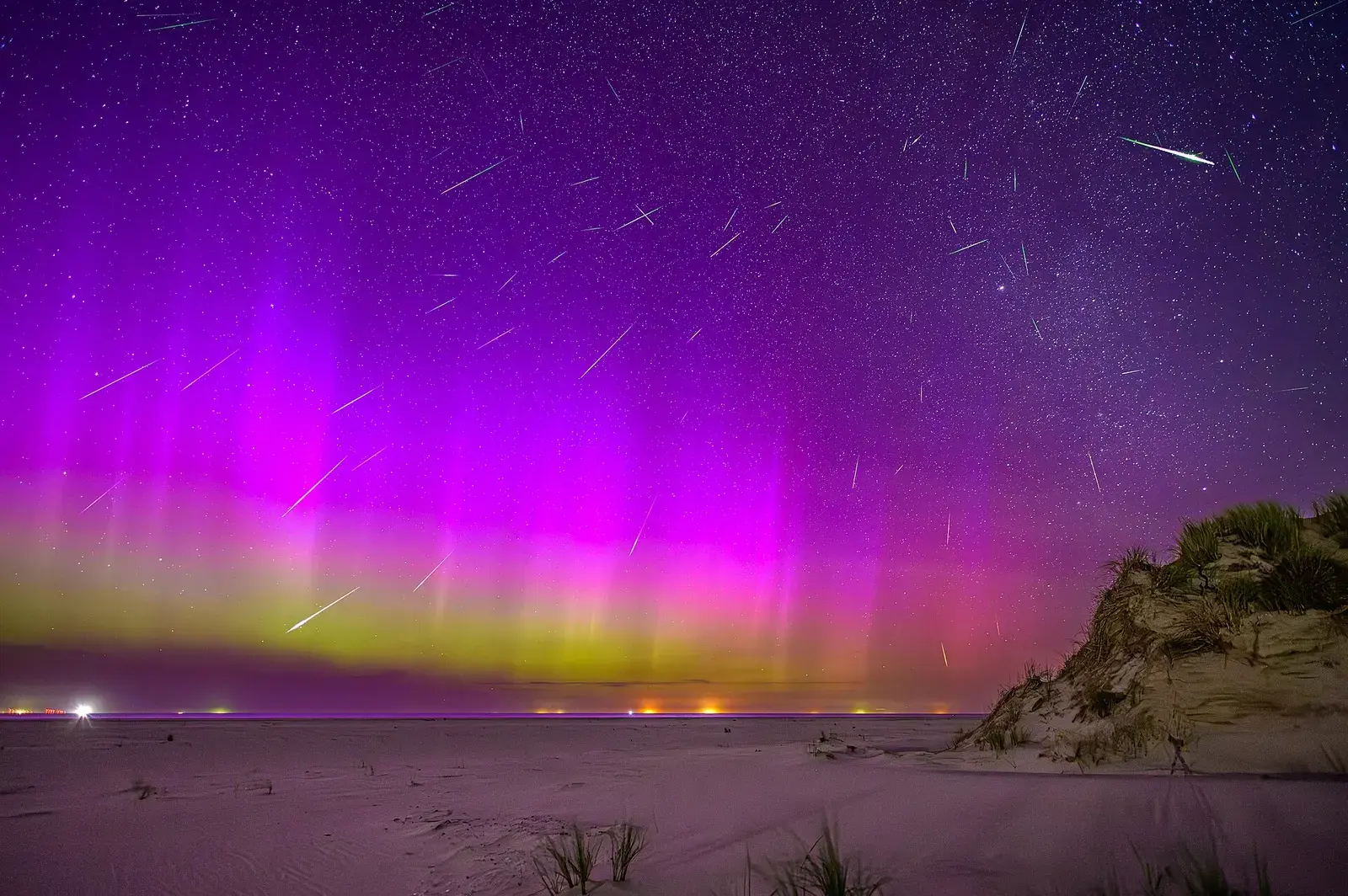 Dozens of meteor streaks across a sky lit pink and green by an aurora, over a beach
