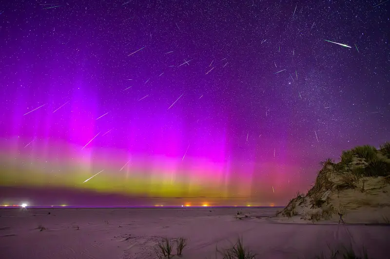 Dozens of meteor streaks across a sky lit pink and green by an aurora, over a beach