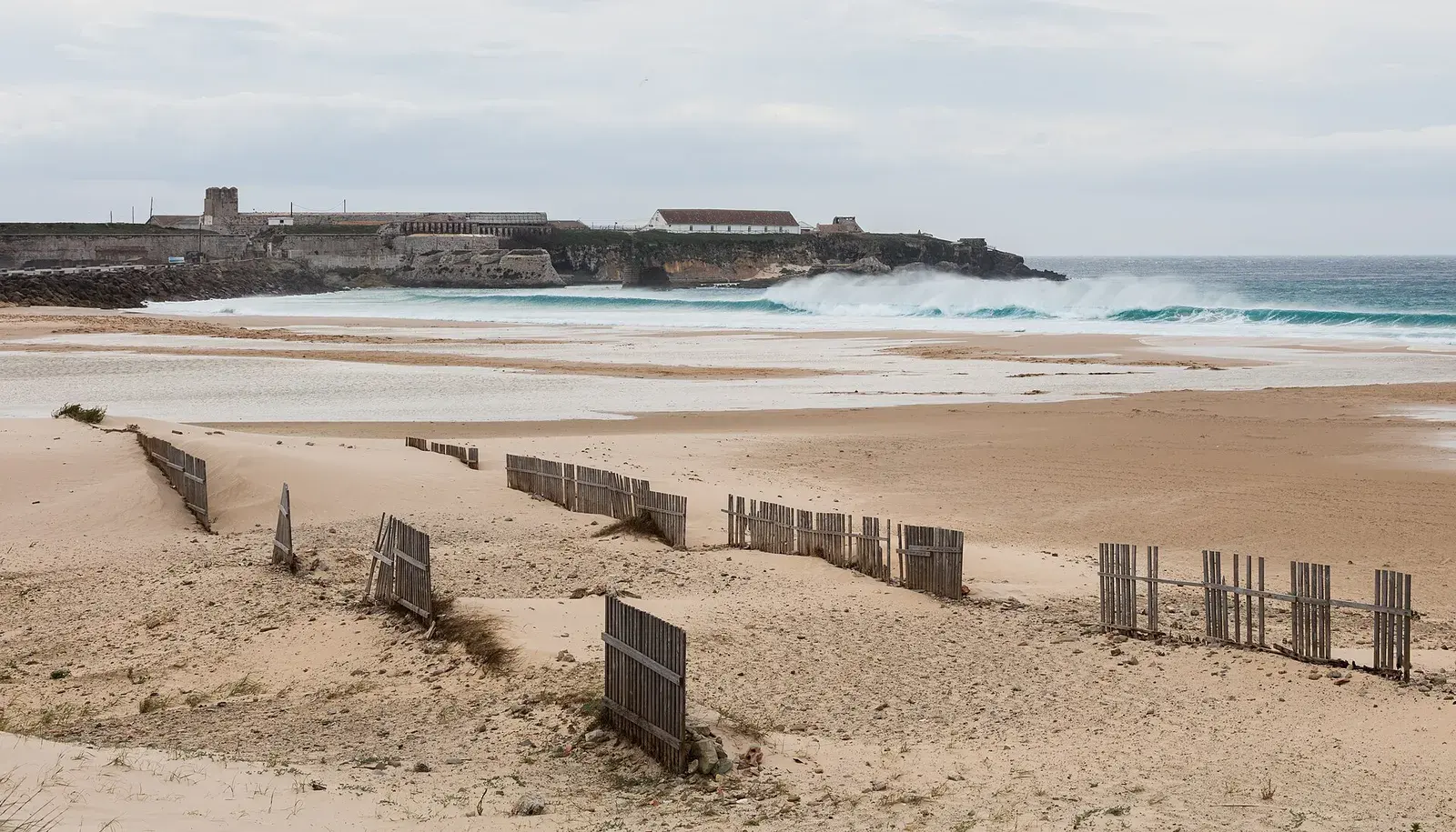 A wide sandy beach with dune fences and waves, with an island fortress behind