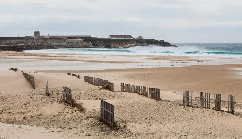 A wide sandy beach with dune fences and waves, with an island fortress behind